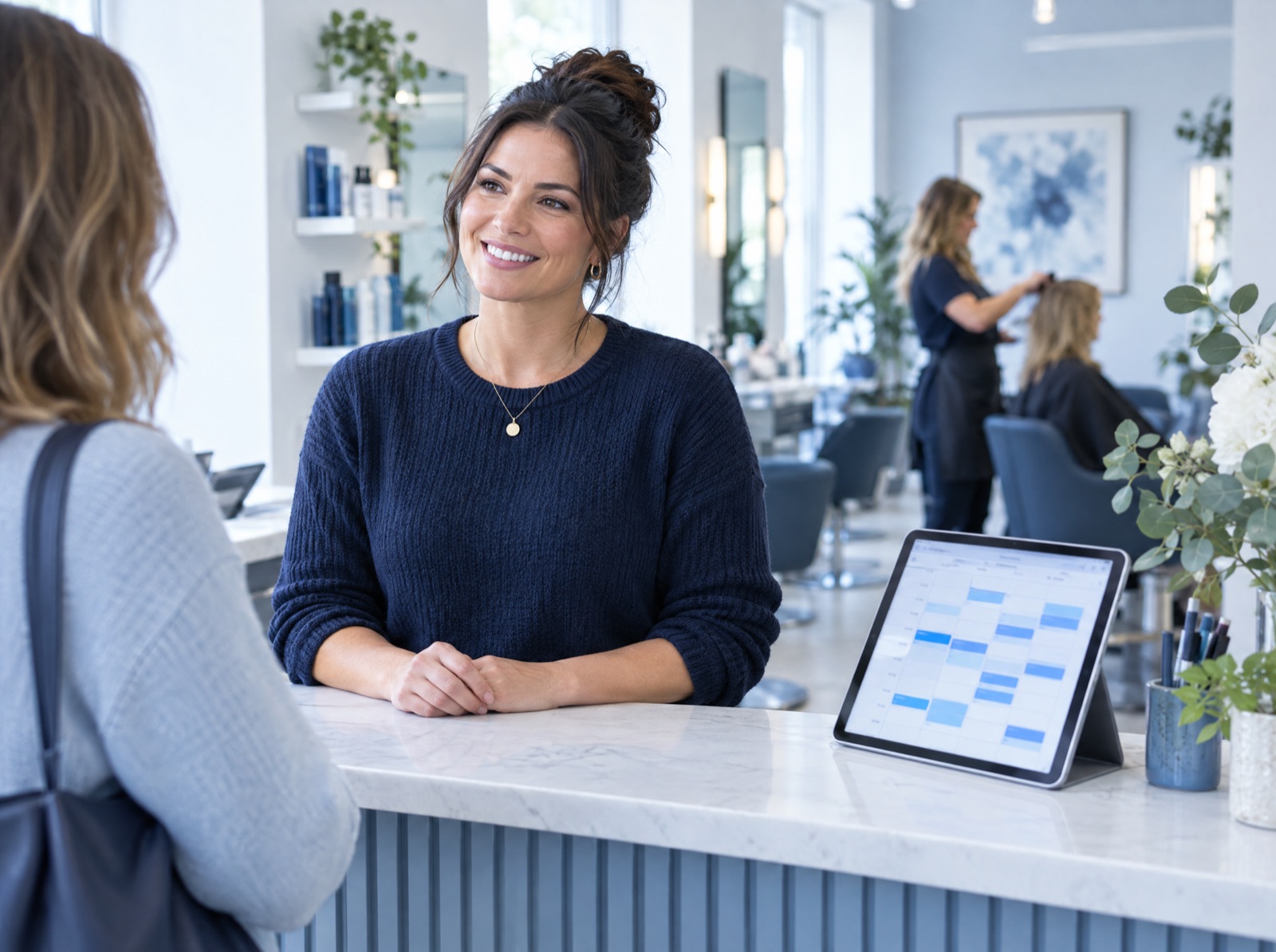 Salon owner chatting with a client at the front desk while a tablet calendar fills with bookings